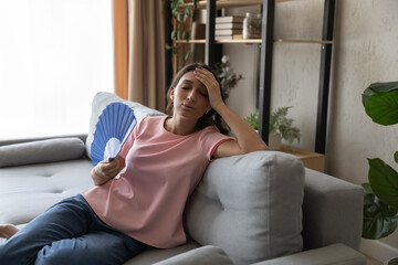 Overheated exhausted Arabian woman waving paper fan, sitting on couch, suffering from hot summer...