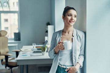 Beautiful young woman in smart casual wear looking away and holding cup