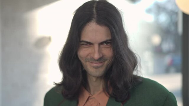 Headshot of confident handsome man with long hair taking off hat winking and putting on headdress. Positive stylish retro guy flirting indoors in 1980s 1990s. Masculinity and confidence