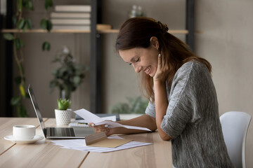 Side view excited young woman reading good news in letter, sitting at desk with laptop, happy...