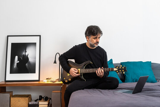 Focused man with laptop playing guitar in bedroom