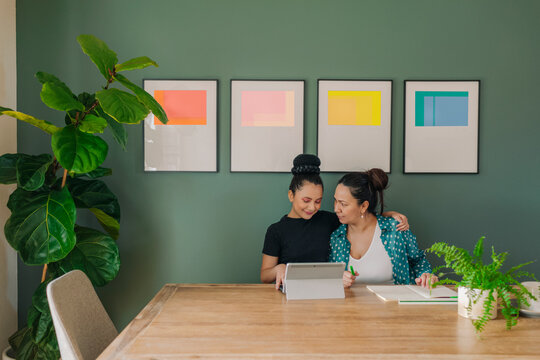 Daughter hugging mother while doing homework in dining table at home by wall gallery
