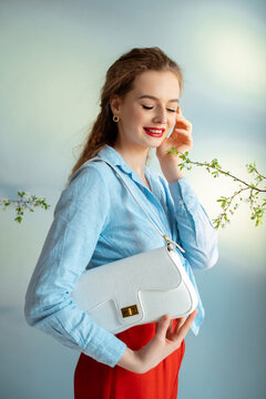 Happy smiling elegant woman wearing blue classic linen shirt, posing with white faux reptile leather textured shoulder bag. Female fashion conception