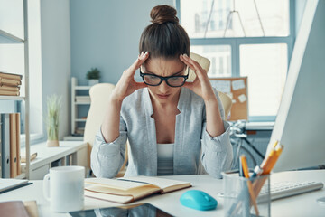 Stressed young woman in smart casual wear keeping head in hands while sitting in the office