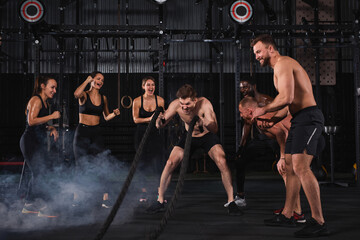 Fototapeta na wymiar Strong shirtless man engaged in cross fit battling ropes at gym workout exercise, while supportive group of friends stand next to him, in sportive outfit