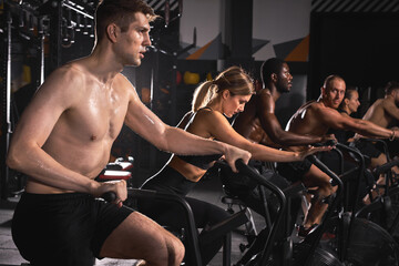 Fototapeta na wymiar Cycling class in fitness club, multi-ethnic group of fit people spinning on cardio machine. Men and women do sports exercises at gym fat burning class. Active lifestyle, health care concept
