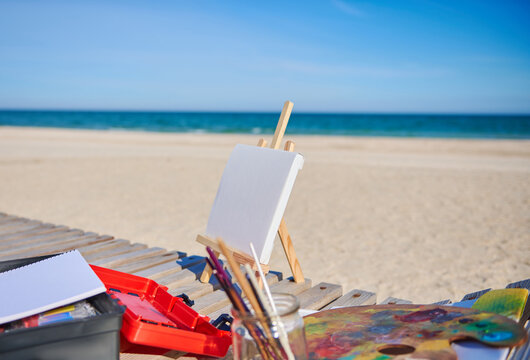 White blank canvas on a wooden table easel next to paints and brushes on a wooden deck chair on the beach against the beautiful blue sea background