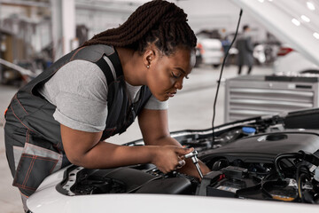 Obraz na płótnie Canvas African woman car mechanic holding wrench checking up on the car engine, for repair and checkup, wearing overall, repairing auto hood. side view portrait. copy space