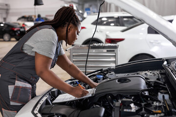 Obraz na płótnie Canvas Young afro american woman mechanic holding clipboard checklist at service center repair, side view on confident serious black female at work, thinking. in auto service
