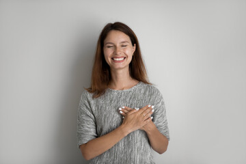 Head shot of smiling woman holding folded hands on chest with closed eyes, standing on grey studio...