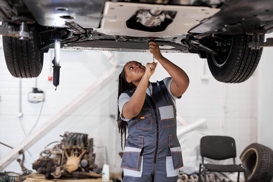 African female auto mechanic fixing car, mechanic repairing car on lift in garage. attractive confident black afro american lady in overalls concentrated on work, repair using instruments tools