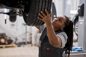 Young afro american mechanic adjusting the tire at repair garage alone, in overalls uniform, black...
