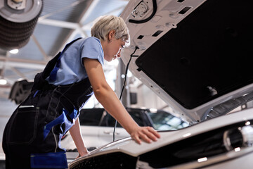 Side View On Pensive Female Auto Mechanic In Overalls Looking Inside Of Car Hood, Woman Stands Thinking, Looking Serious And Confident, In Overalls. In Auto Service