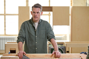Portrait of confident caucasian male carpenter in green shirt polishing wooden board with...