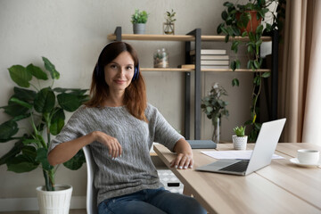 Portrait of confident woman in headphones sitting at desk with laptop, studying online, listening to lecture, working on project, attractive young female student or freelancer looking at camera