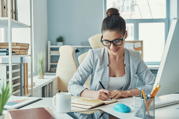 Confident young woman in smart casual wear writing something and smiling while sitting in the office