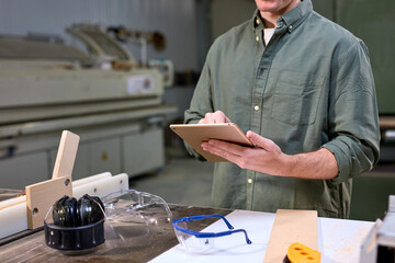 Cropped carpenter in green shirt using digital table at work standing next to table with tools and wooden materials. side view portrait of man joiner in workshop, alone. copy space