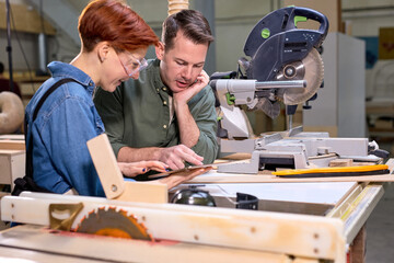 Obraz premium Customer and joiner have talk in workshop, discussing, sharing ideas what to make from wood, handmate furniture. two caucasian man and woman indoors, sawing machine in the background