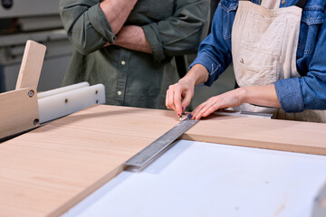 hardworking focused professional serious carpenter woman holding ruler and pencil while making marks on wood at table in fabric workshop, concentrated on work, making furniture. close-up hands