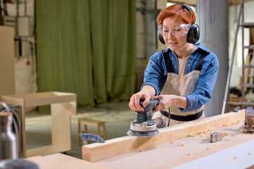 Young female carpenter working with wood using electric sander in workshop. Joinery work on the production and renovation of wooden furniture. Small Business Concept