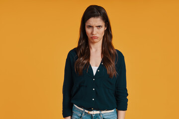 Sad young woman in casual clothing making a face while standing against yellow background