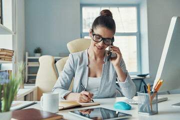 Beautiful young woman in smart casual wear talking on the phone and smiling while sitting in the office