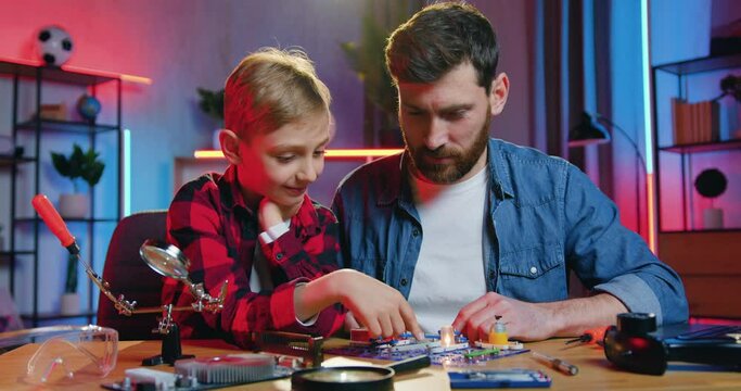 Pleasant high-skilled smiling 35-aged bearded man collecting small details of constructor to light the lamp and curious teen son is surprised from seen result