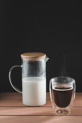 Black coffee in glass cup with double walls. Milk decanter. Wooden table and black table.