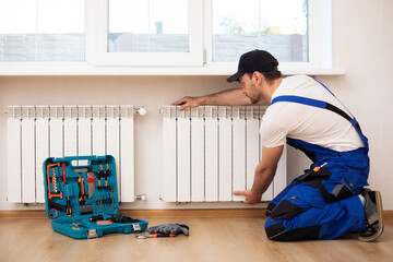 Professional young repairman in special uniform with tools is installing radiators and thermostat...