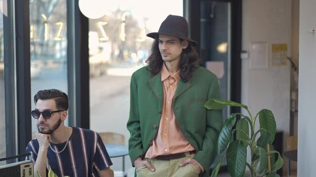 Confident 1980s dandy walking in slow motion looking at people sitting in vintage coffee shop. Portrait of masculine Caucasian man in hat and velvet jacket strolling indoors. 1990s style concept