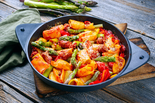Traditional roasted Italian pasta mezzi paccheri rigati con salsiccia sausage with green asparagus tips in tomato sauce served as close-up in cast iron skillet pan in matte flat cinematic modern look