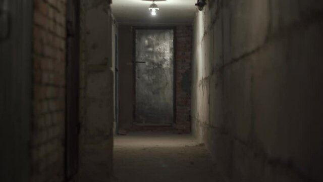 First person view moving straight down a narrow empty dark narrow corridor with concrete walls under the ground with dim light. Underground catacombs of World War II. Metal door at the end of corridor