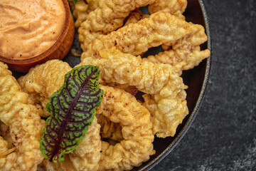 Golden rings of squid fries in a round frying pan with sauce, herbs and lemon on a dark background without decorations