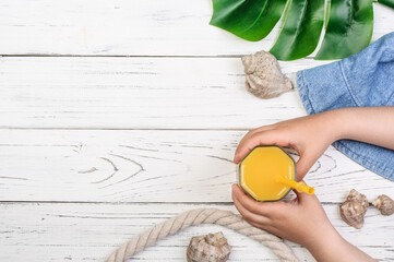 child holding glass with juice, near seashells, monstera leaf and hat on white boards