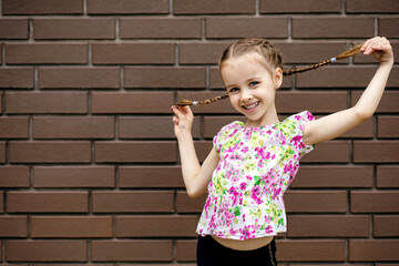 A little girl stands against a brick wall and playfully holds her pigtails. Cute baby shows...