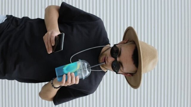 Vertical shot of stylish man drinking tropical cocktail and using smartphone. Handsome male hipster drinking cold refreshing summer beverage and browsing mobile phone in city