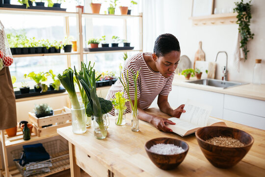 Black woman reading book near growing vegetables in kitchen
