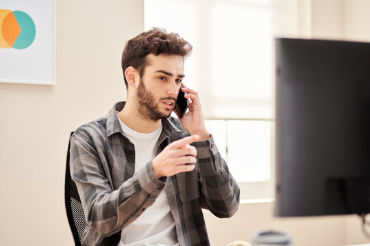 Focused freelancer speaking on phone while using desktop computer
