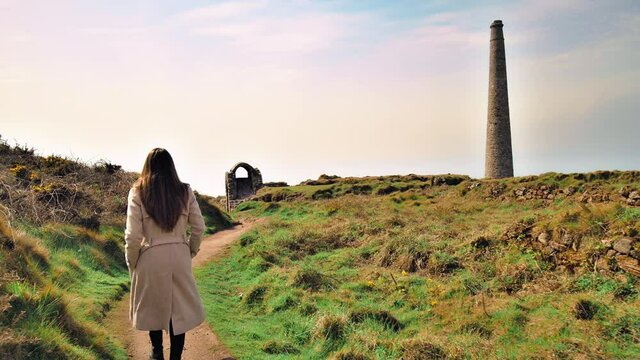 A fashionable girl in a coat walking through ruins of a castle in Cornwall in 4K. Crowns Engine Houses, Botallack Mine, ruins of an old ancient English building in 4k.
