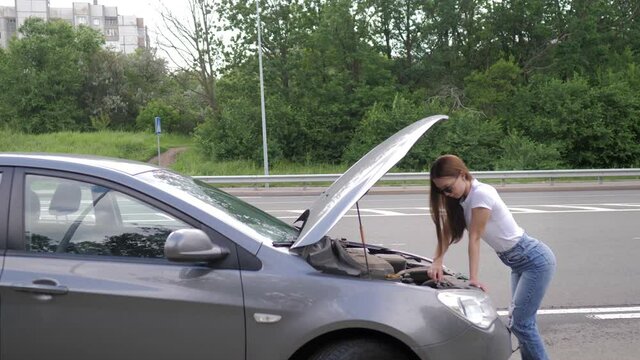 A young beautiful girl stands by a wrecked car on the highway. A girl with glasses looks under the hood of a broken car, trying to fix it. Problems with the car. A girl on the road repairs a car