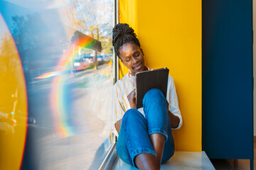 Black woman browsing tablet sitting on windowsill