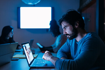 Serious man working on laptop in office with diverse colleagues at night