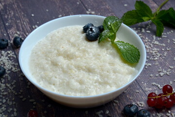 Rice coconut porridge with berries and coconut milk in bowl on rustic wooden background. Healthy breakfast ingredients. Clean eating, vegan food concept, close up