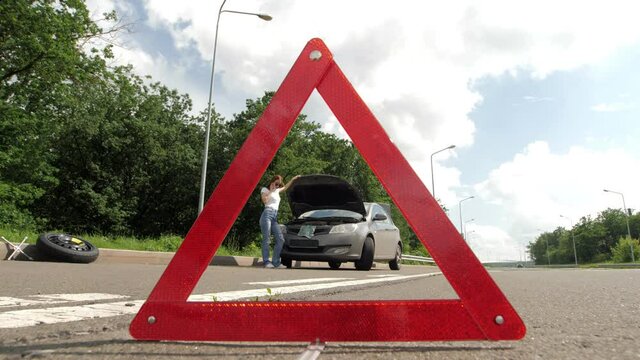 A young beautiful girl stands by a broken car on the highway on a sunny day and calls the car service. The girl is standing near the open hood. Repair of cars on the highway. Emergency stop sign