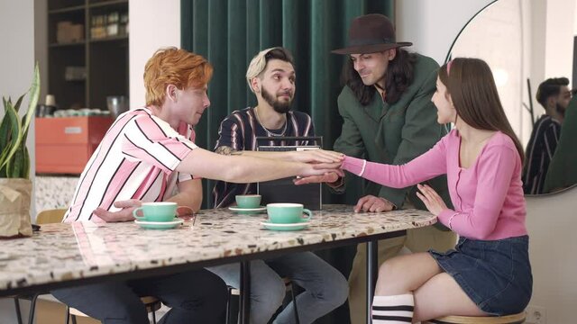 Young friends stacking hands in retro coffee bar smiling. Group of positive Caucasian men and women resting indoors in 1980s cafe. Friendship and vintage concept