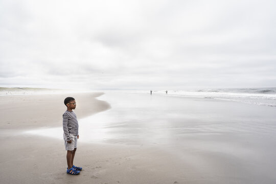 Black boy stands looking out into the ocean on cloudy day 