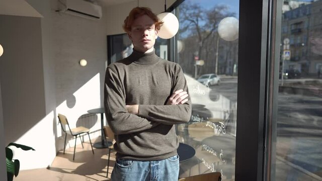 Confident redhead man crossing hands in slow motion looking at camera standing in cafe indoors. Portrait of handsome Caucasian guy posing in coffee shop. Lifestyle and confidence