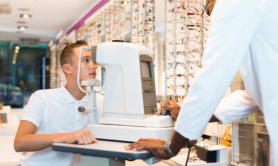 Teenage boy receiving eye examination on modern refractometer in optical store with professional serious African-American ophthalmologist - obrazy, fototapety, plakaty