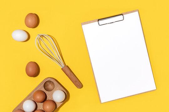 Eggs in wooden egg box, whisk and clipboard with white paper on yellow background. Flat lay Top view Mockup