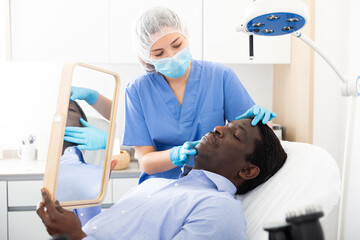 Fototapeta premium Skilled female beautician in face mask giving consultation to african male patient in aesthetic clinic